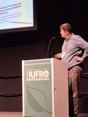 A male researcher standing in front of a lecturn with the word IUFRO on a green background with a leaf design.